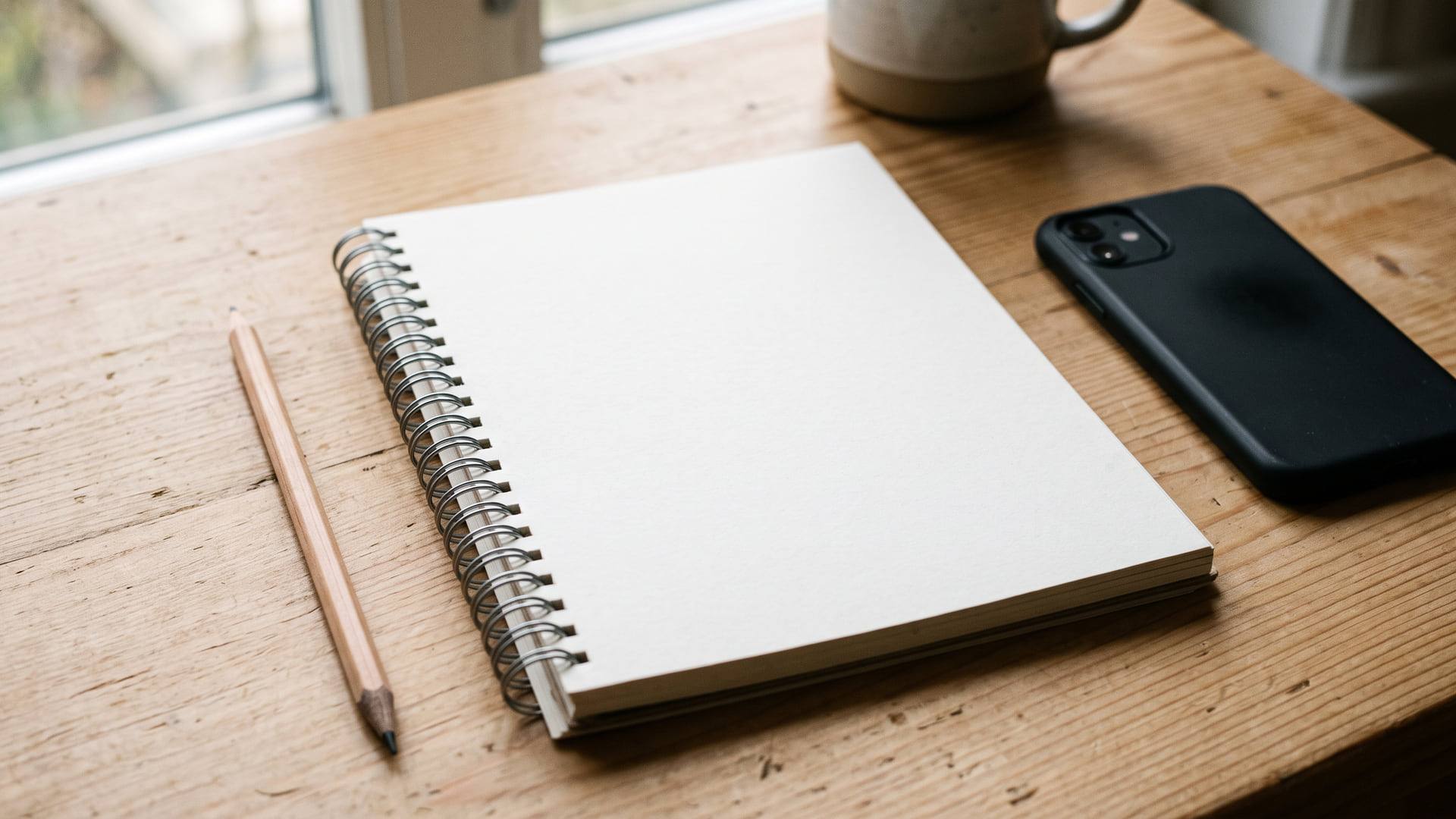 Carnet à spirale ouvert sur une page vierge, crayon et smartphone retourné posés sur une table en bois clair