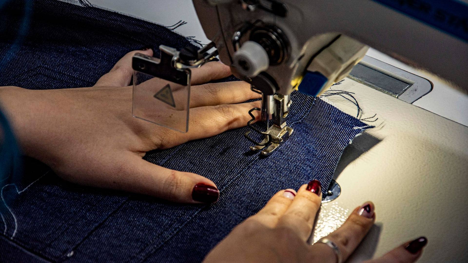 Une personne retravaillant un vêtement en denim usagé à l'aide d'une machine à coudre moderne sur une table en bois.